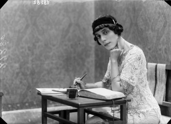 Una fotografía en blanco y negro en la que se ve a Amalia Guglielminetti sentada frente a una pequeña mesa. Tiene un libro delante de ella y sostiene una pluma en la mano derecha. Tiene puesto un vestido claro de encaje o una tela similar y un sombrero pequeño y de color oscuro. Su cabello es corto, enrulado y oscuro. Ella mira a la cámara sin sonreír y tiene el mentón apoyado sobre el puño izquierdo. Parece que estaba escribiendo y alguien hizo que alzara la para retratarla
