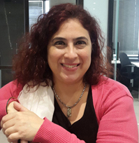 alt text:En un primer plano, Ana Saladino mira directamente a cámara y sonríe. Su cabello es castaño rojizo y con rulos. Tiene puesta una remera negra y un suéter rosa.