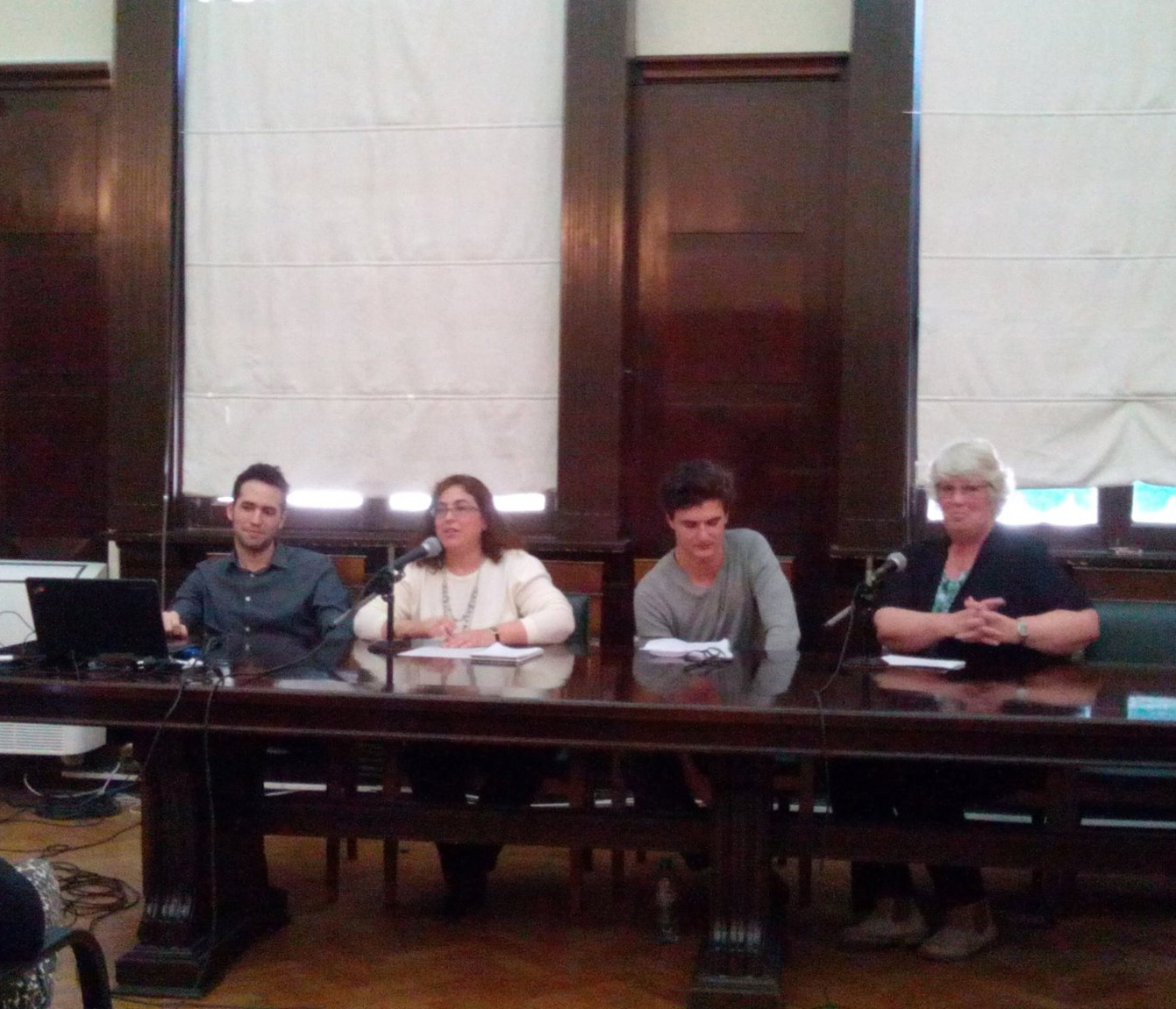 En una sala de la facultad de derecho, Ana Saladino está sentada frente a una mesa en compañía de tres personas. Ana está hablando a un micrófono.