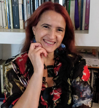 alt text:Sobre el fondo de una biblioteca blanca, se ve un primer plano de Erika Cosenza mirando a cámara y sonriendo. Está vestida con una camisa en tonos rojos y negros. Su cabello es largo, lacio y rojo.