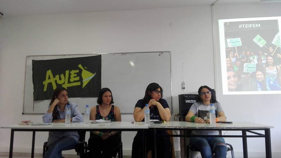 La fotografía muestra un aula de facultad. Hay una mesa con cuatro mujeres, entre las cuales se encuentra Erika Cosenza. En la pared del fondo se ve una pancarta con el nombre del centro de estudiantes, AULE.